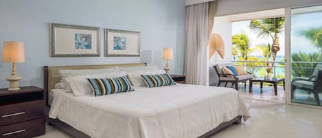A hotel room with a bed, two bedside lamps, a window with a view of palm trees, and a balcony with outdoor furniture.