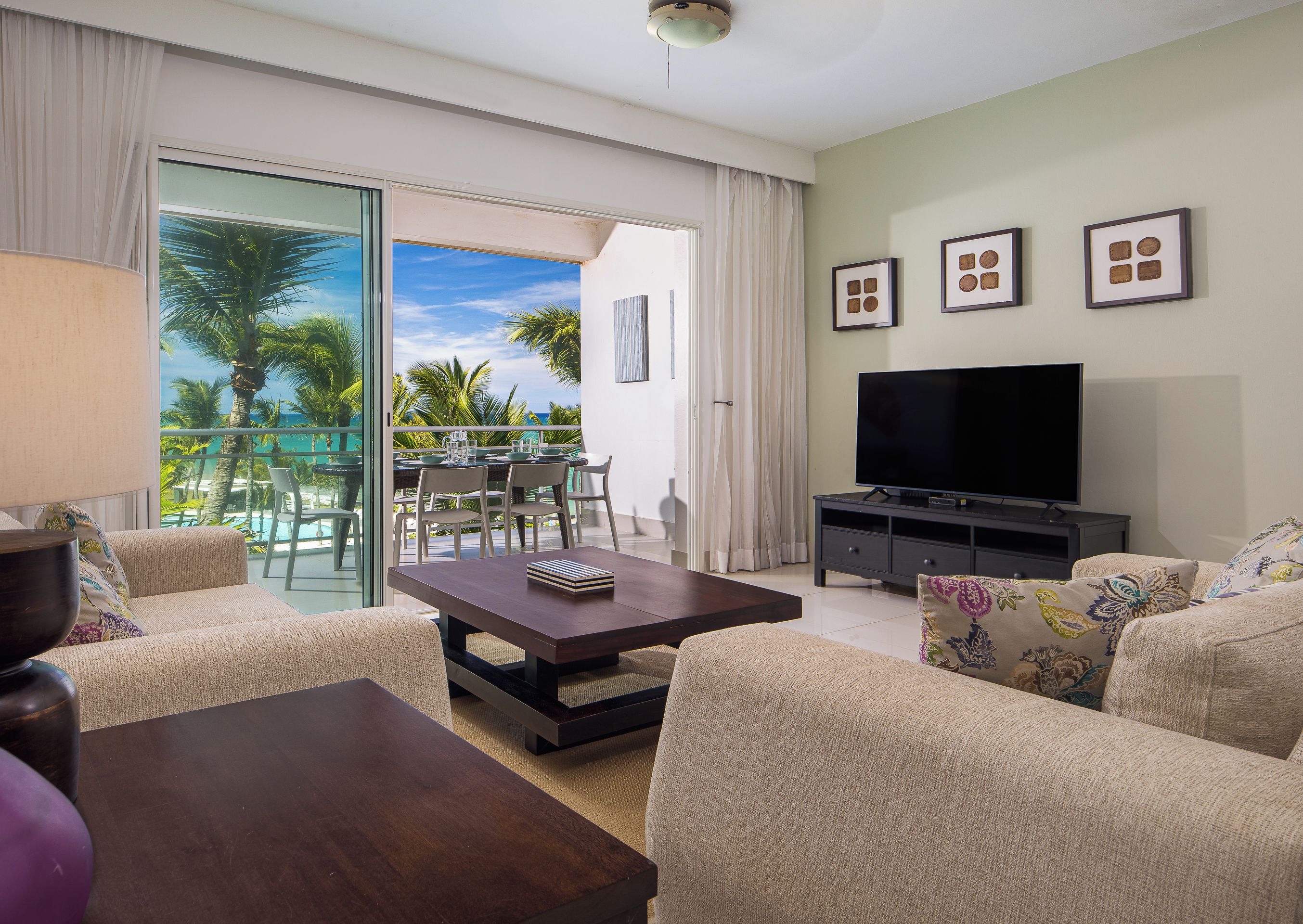 A living room with a TV, sofa, coffee table, and a balcony with a view of palm trees and the ocean.