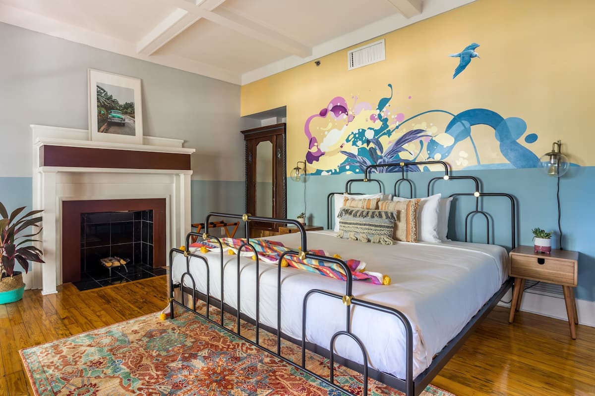 Een slaapkamer met een metalen bedframe, een open haard, een kleurrijk tapijt en een muurschildering.