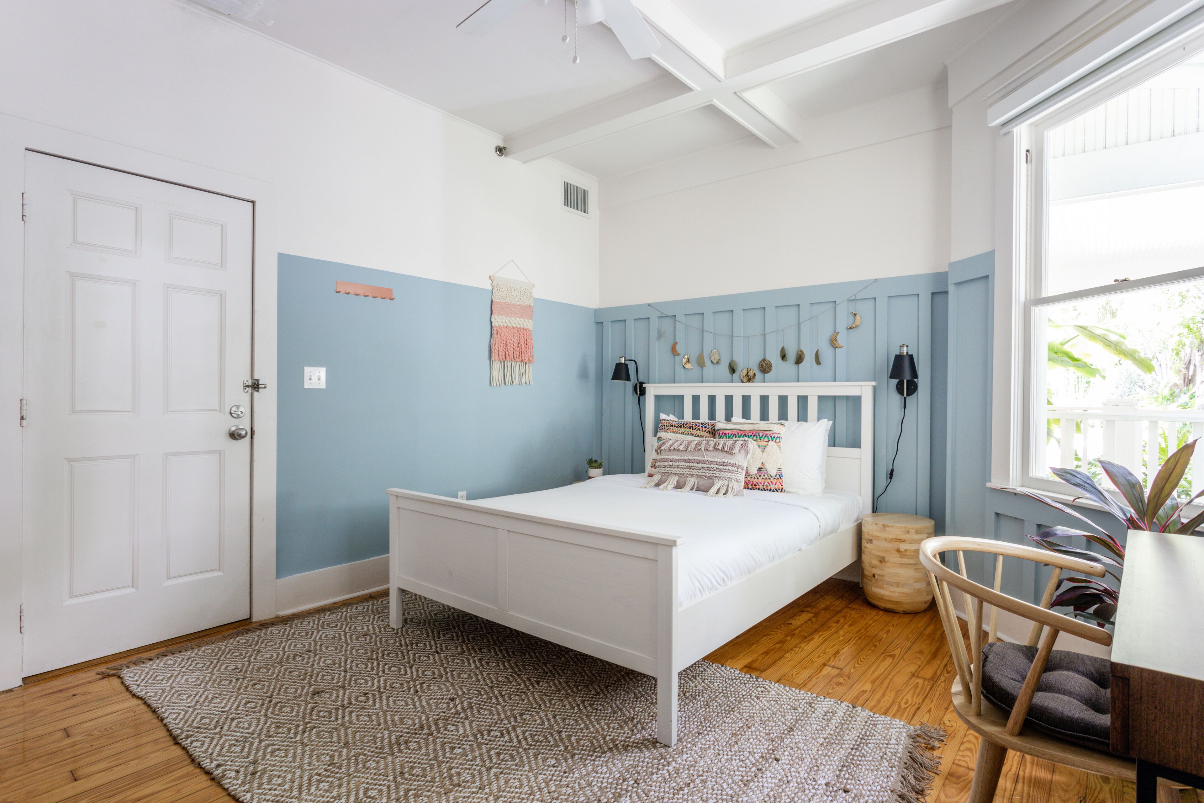 A bedroom with a white bed, blue walls, a wooden floor, a patterned rug, and a window with a view of greenery.