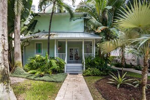A two-story house with a porch, surrounded by tropical plants and a pathway leading to the entrance.