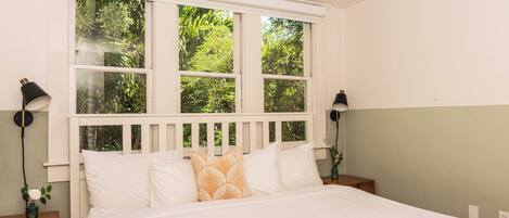 A bedroom with a large bed, white headboard, and a window with a view of greenery. There are two bedside tables with lamps and a small plant on one.