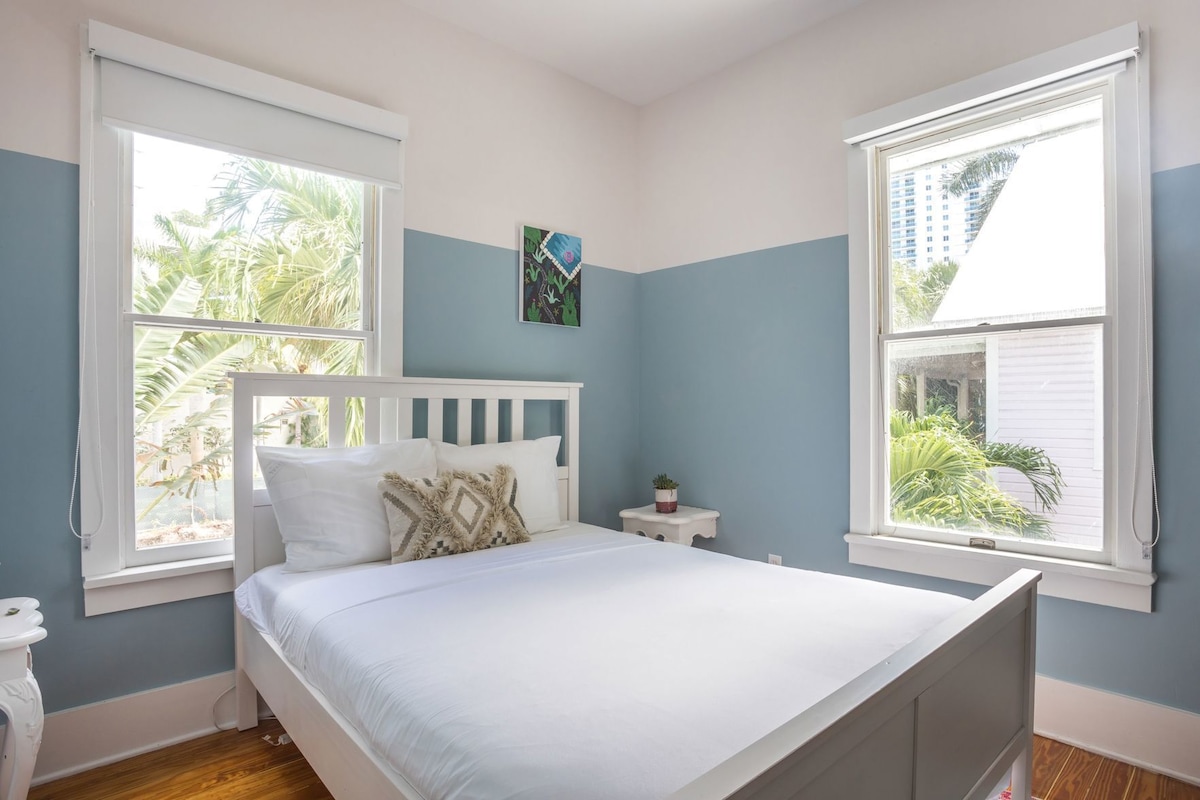 A bedroom with a bed, two windows, a nightstand, a small table, and a wall-mounted artwork.