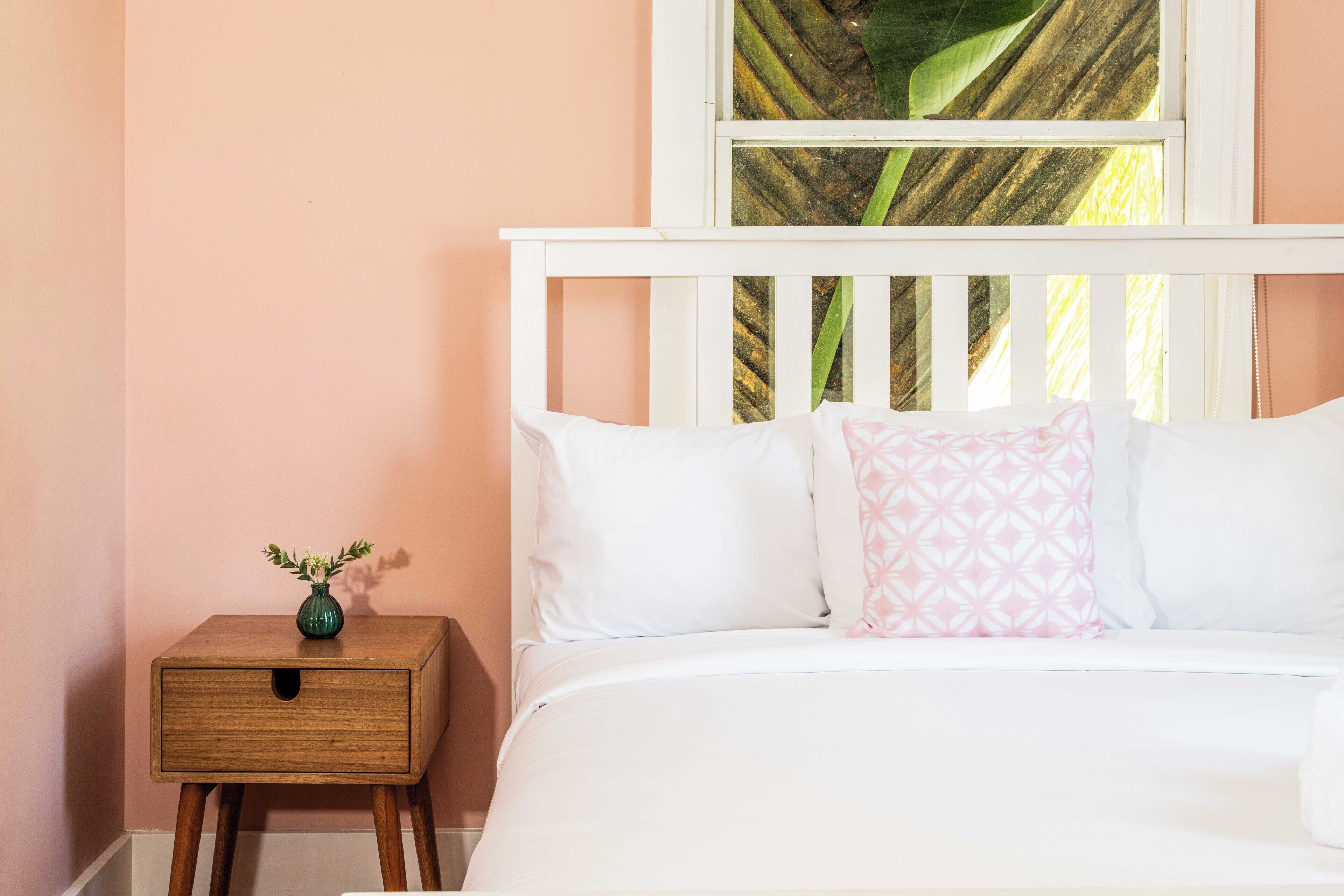 Un dormitorio con una cama blanca, una mesita de noche de madera con un jarrón y una ventana con vistas a zonas verdes.