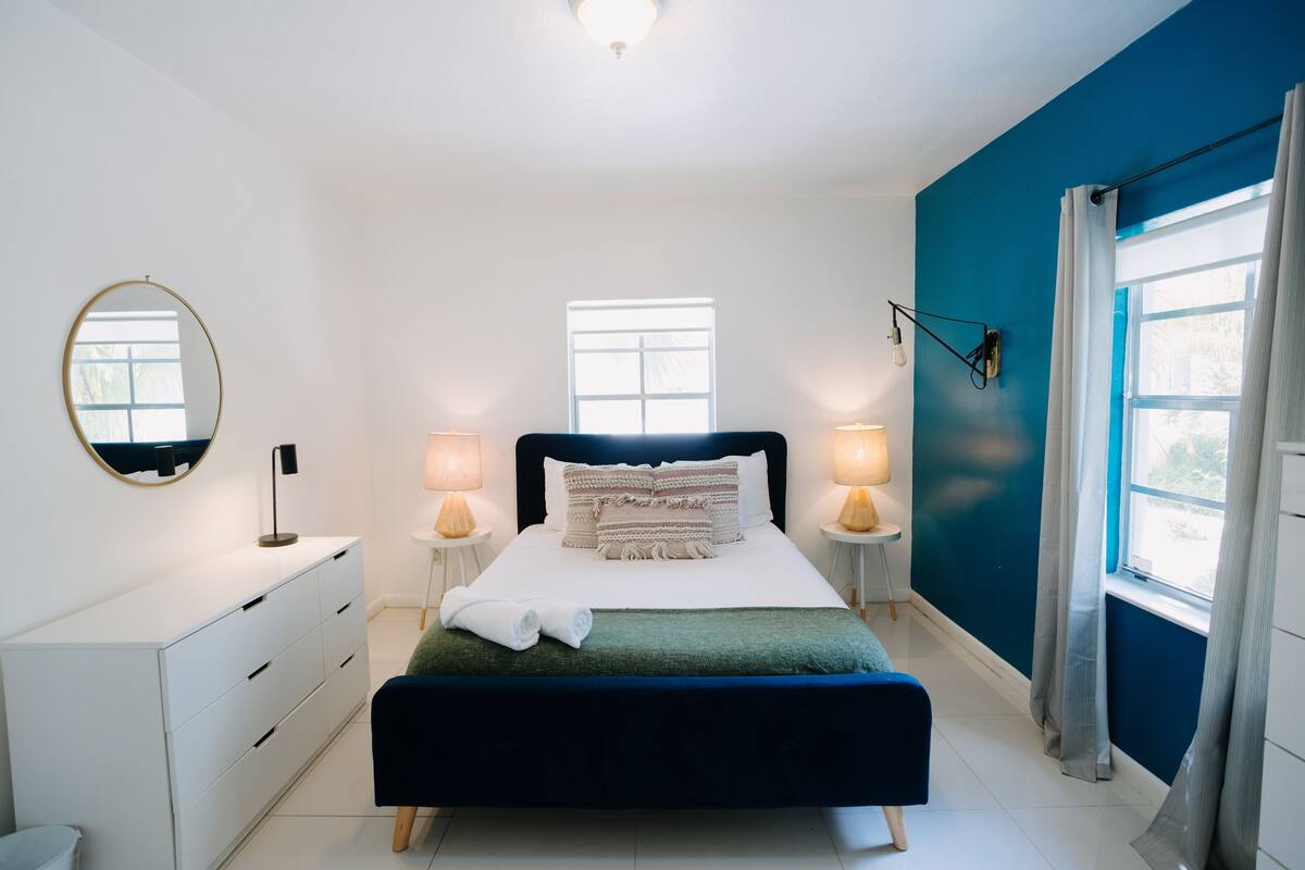 Een slaapkamer met een bed, nachtkastjes, een dressoir, een spiegel en een raam met gordijnen.