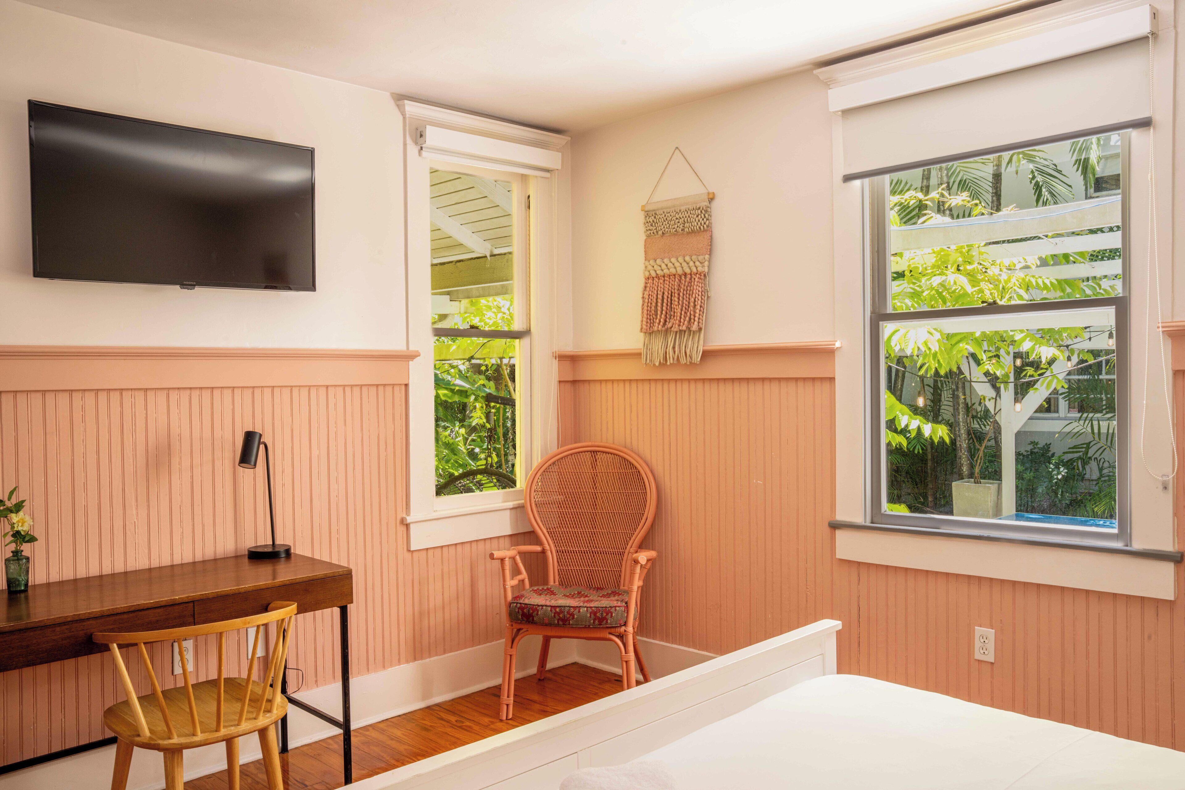 A room with a wooden desk, a wicker chair, a bed, a TV mounted on the wall, and a window with a view of greenery.