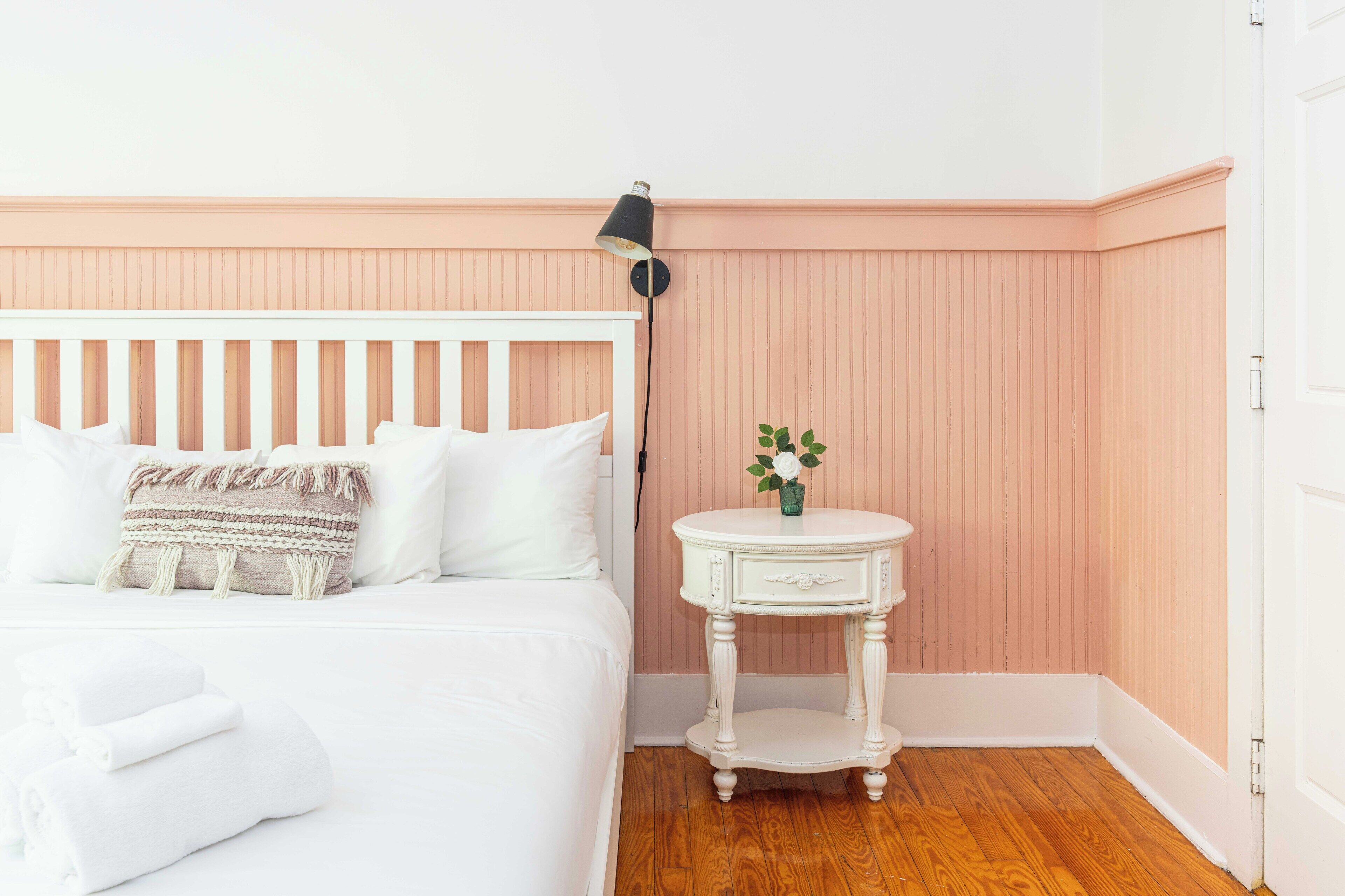 Uma cama bem arrumada com roupa de cama branca e uma almofada decorativa, uma pequena mesa redonda com um vaso de flores, e um abajur fixado na parede.