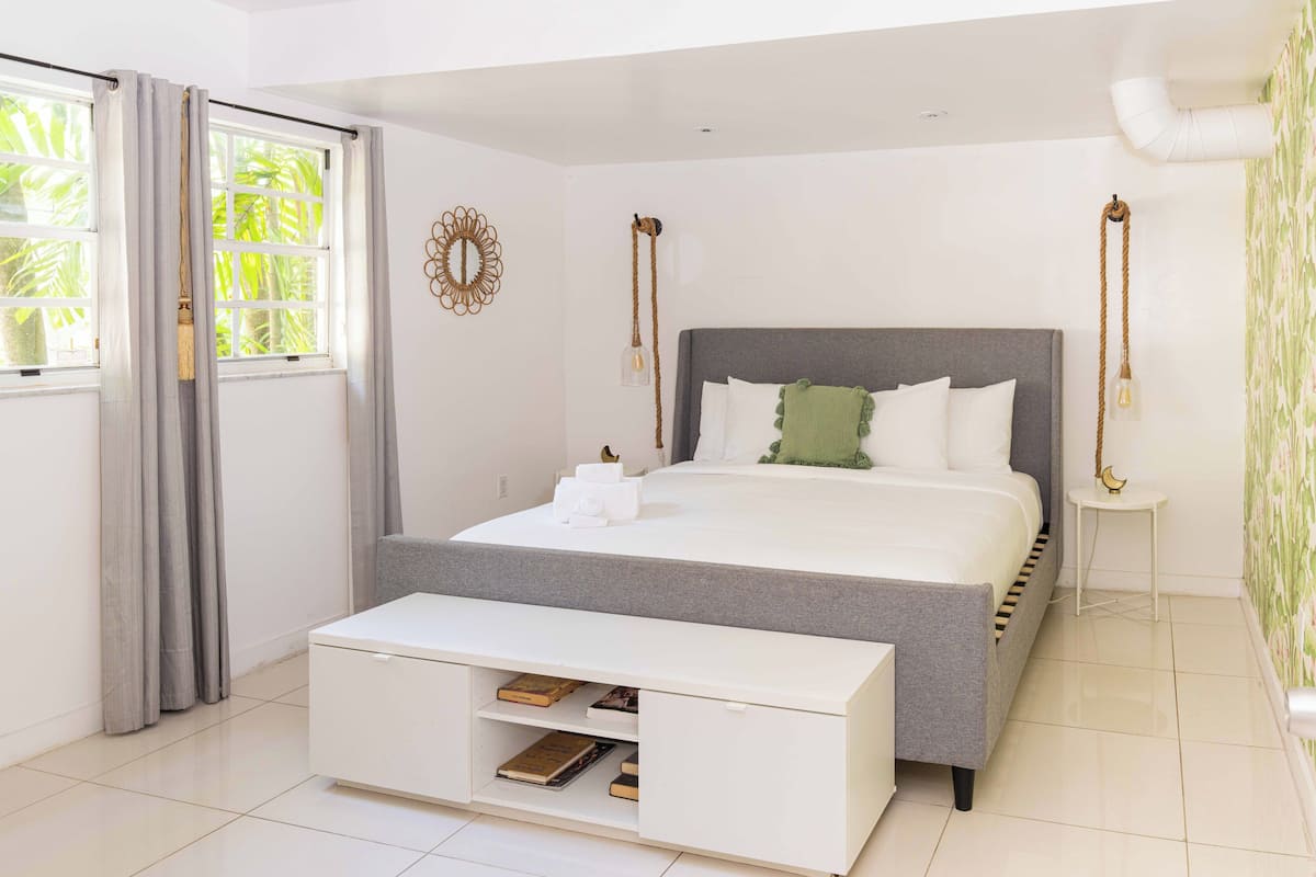 Een moderne slaapkamer met een grijs bed, witte nachtkastjes en een witte console met boeken.