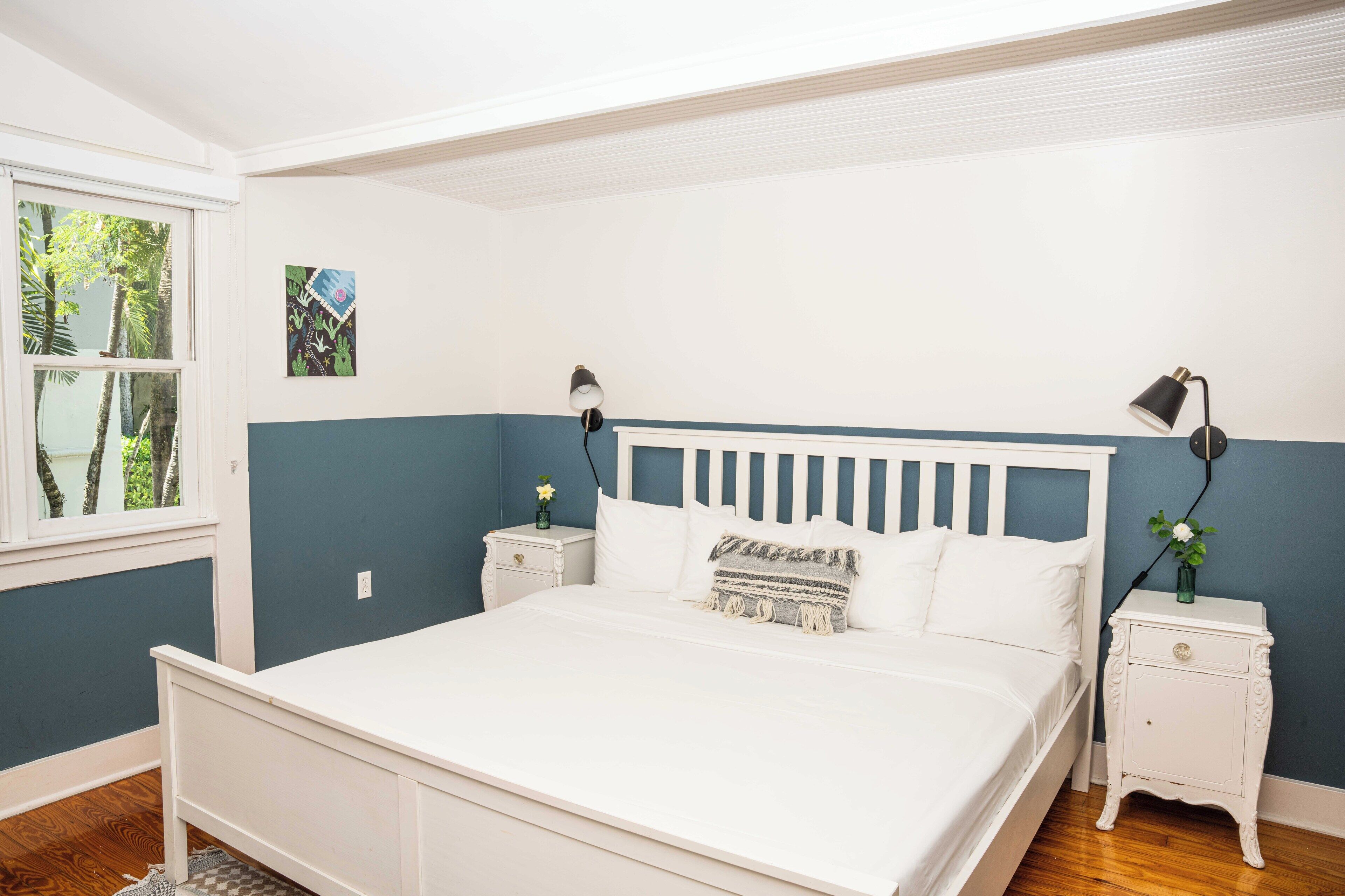 A bedroom with a white bed, two nightstands, a window with a view of trees, and a wall with a blue accent.