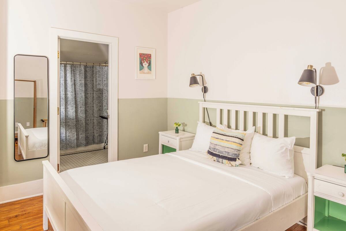 Een slaapkamer met een bed, nachtkastjes, een spiegel, een foto aan de muur en een deur naar de badkamer.
