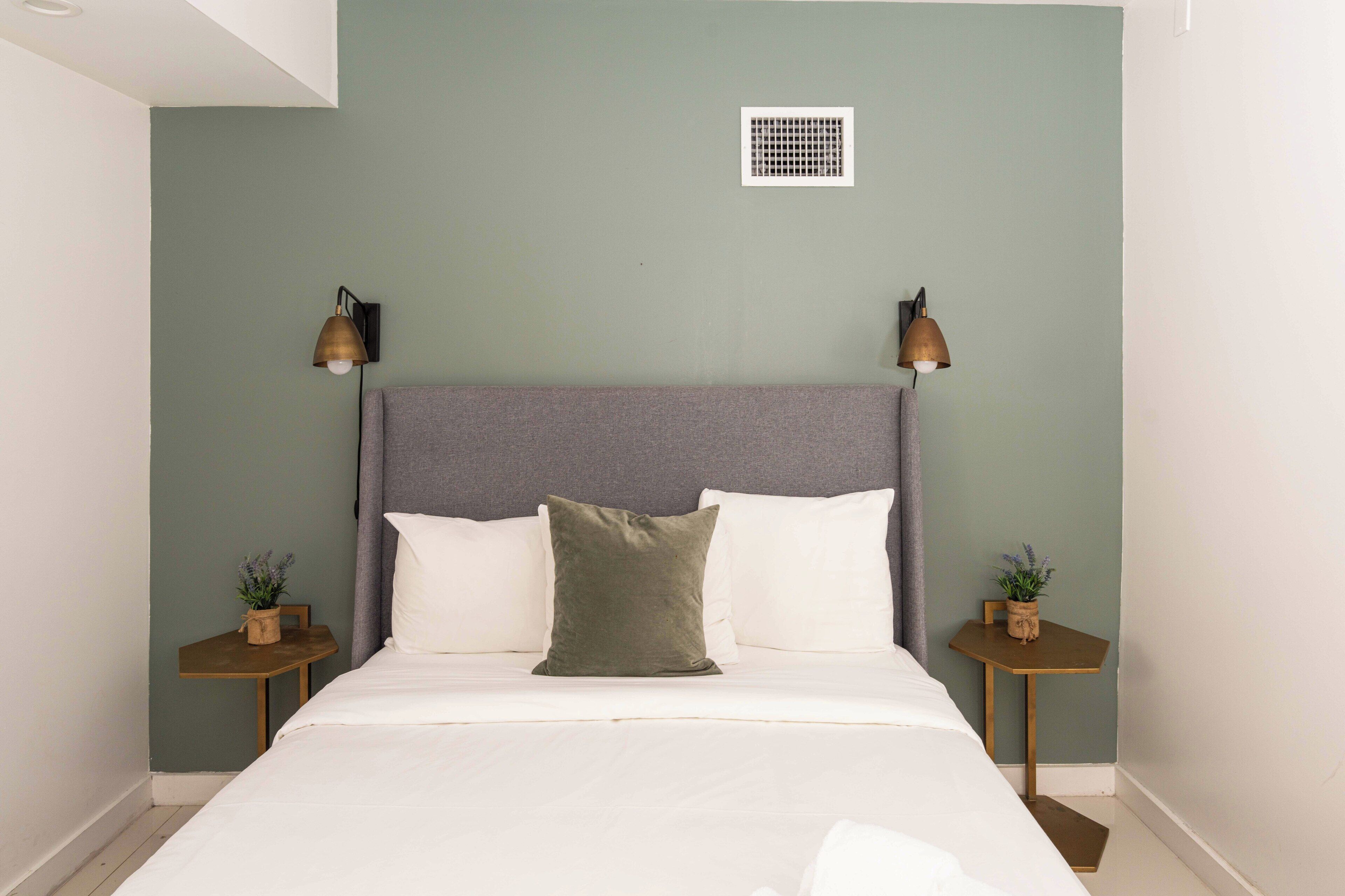 Una cama bien tendida con ropa blanca y un almohadón verde, flanqueada por dos mesitas de noche con pequeñas plantas en macetas.