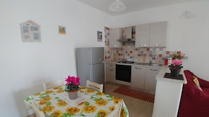 A compact kitchen with a refrigerator, stove, and sink. There's a dining table with a floral tablecloth and chairs. A red sofa is in the corner.