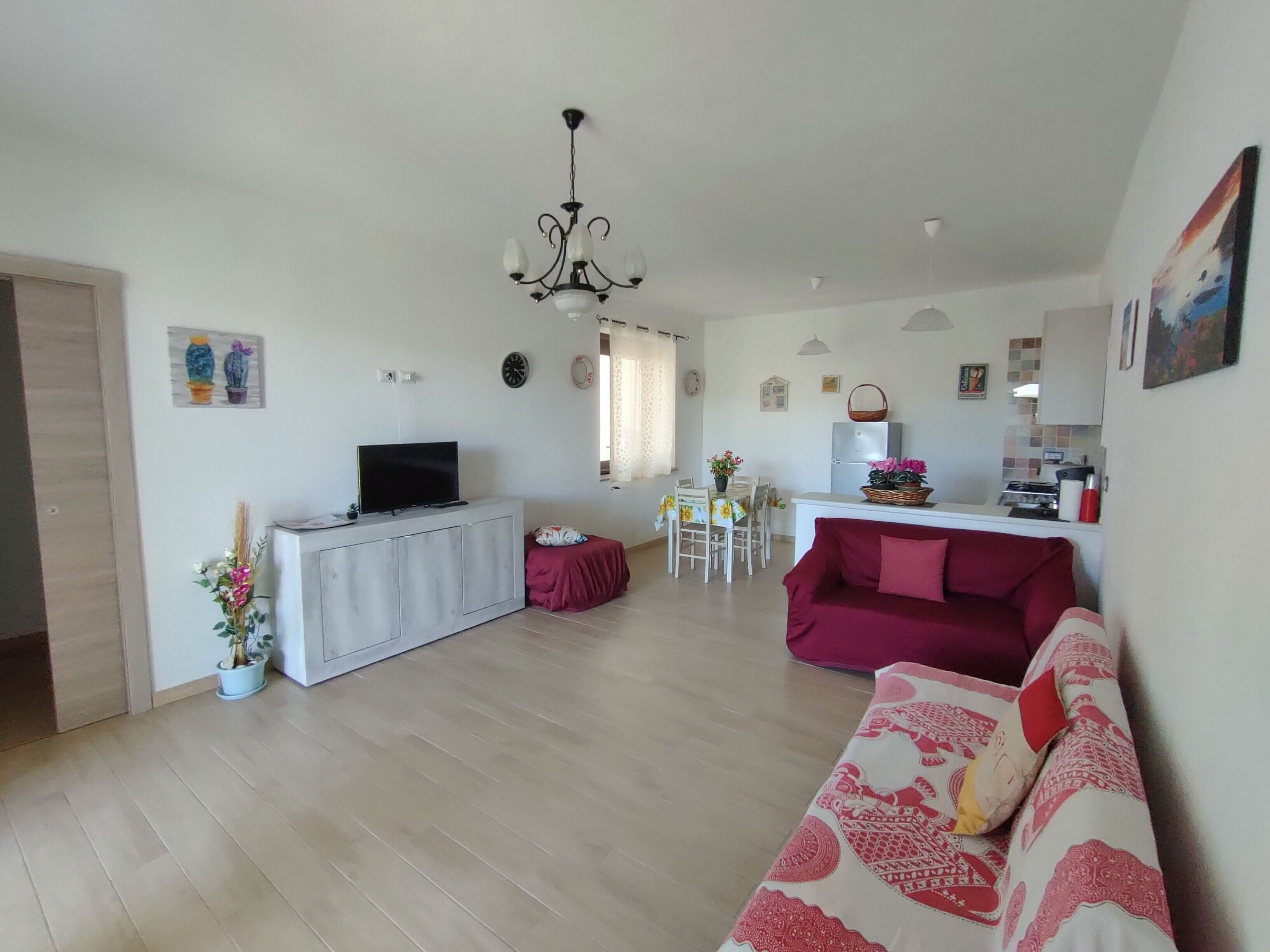 A living room with a TV, a red sofa, a dining area with a table and chairs, and a kitchen area with a fridge and stove.