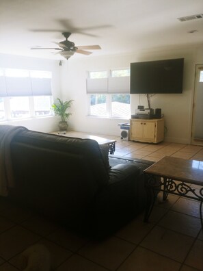 A living room with a sofa, a coffee table, a ceiling fan, a television, and a window with blinds.