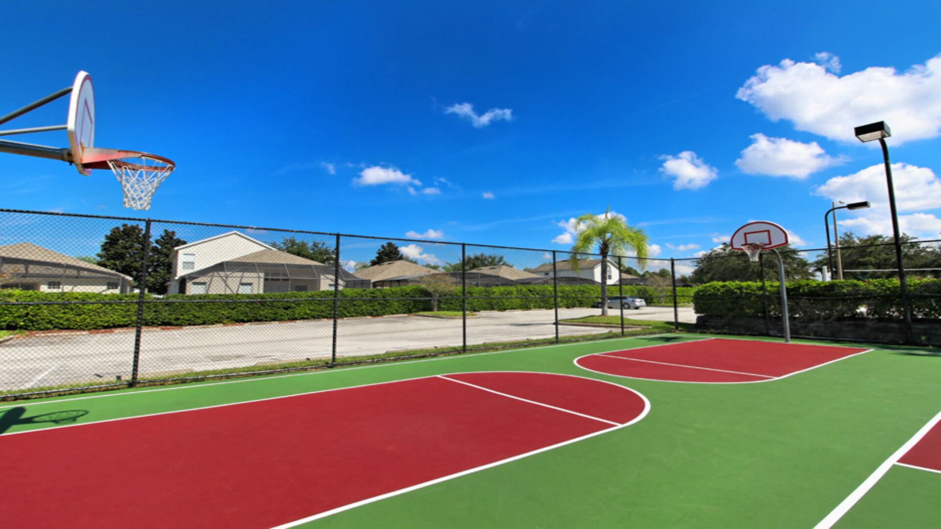Una cancha de baloncesto al aire libre con dos aros, rodeada por una valla, y casas al fondo.