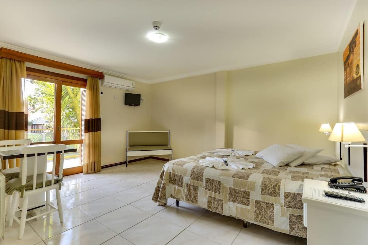 Quarto de hotel com cama, escrivaninha, cadeira, televisão e janela com cortinas.