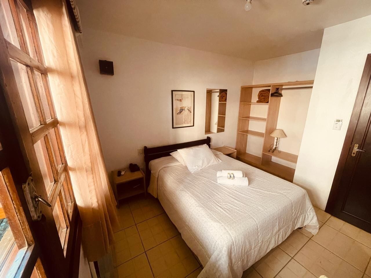 A neatly made bed with white linens, a wooden headboard, a nightstand with a lamp, and a wall-mounted shelf with a lamp.