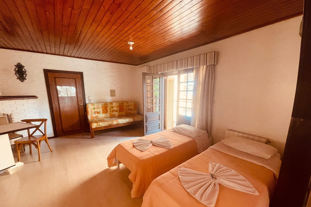 Quarto com duas camas, uma escrivaninha, uma cadeira, um banco e uma janela com cortinas.