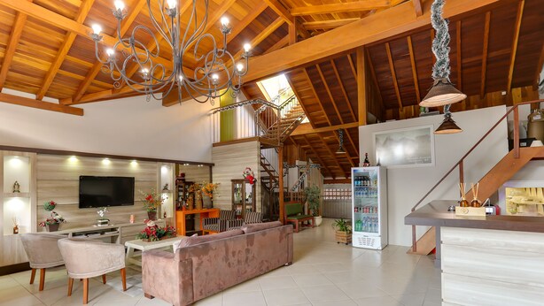 A spacious living area with a high ceiling, wooden beams, a large chandelier, a flat-screen TV, a sofa, chairs, a coffee table, and a refrigerator. - Zermatt Hotel (Gramado)