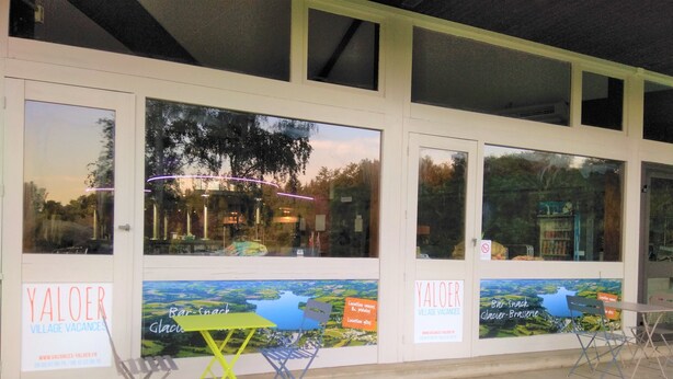 A Yaloer Village Vacances storefront with large windows, a bar-snack-glacier-brasserie sign, and outdoor seating with chairs and a table.
