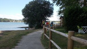 A gravel path leading to a wooden fence by a lake, with a small building and green chairs in the background. - Le Camping St Etienne (Villefranche-de-Panat)
