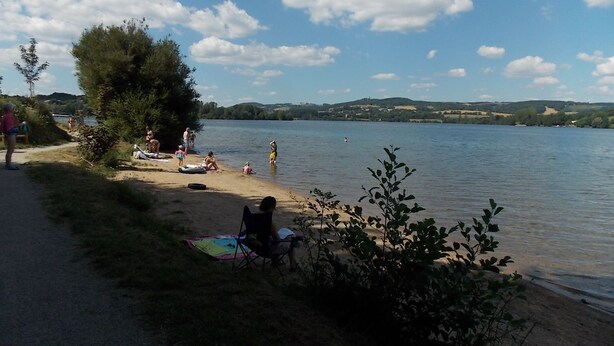 A lakeside setting with a sandy beach, a few people, and a clear sky. - Le Camping St Etienne (Villefranche-de-Panat)