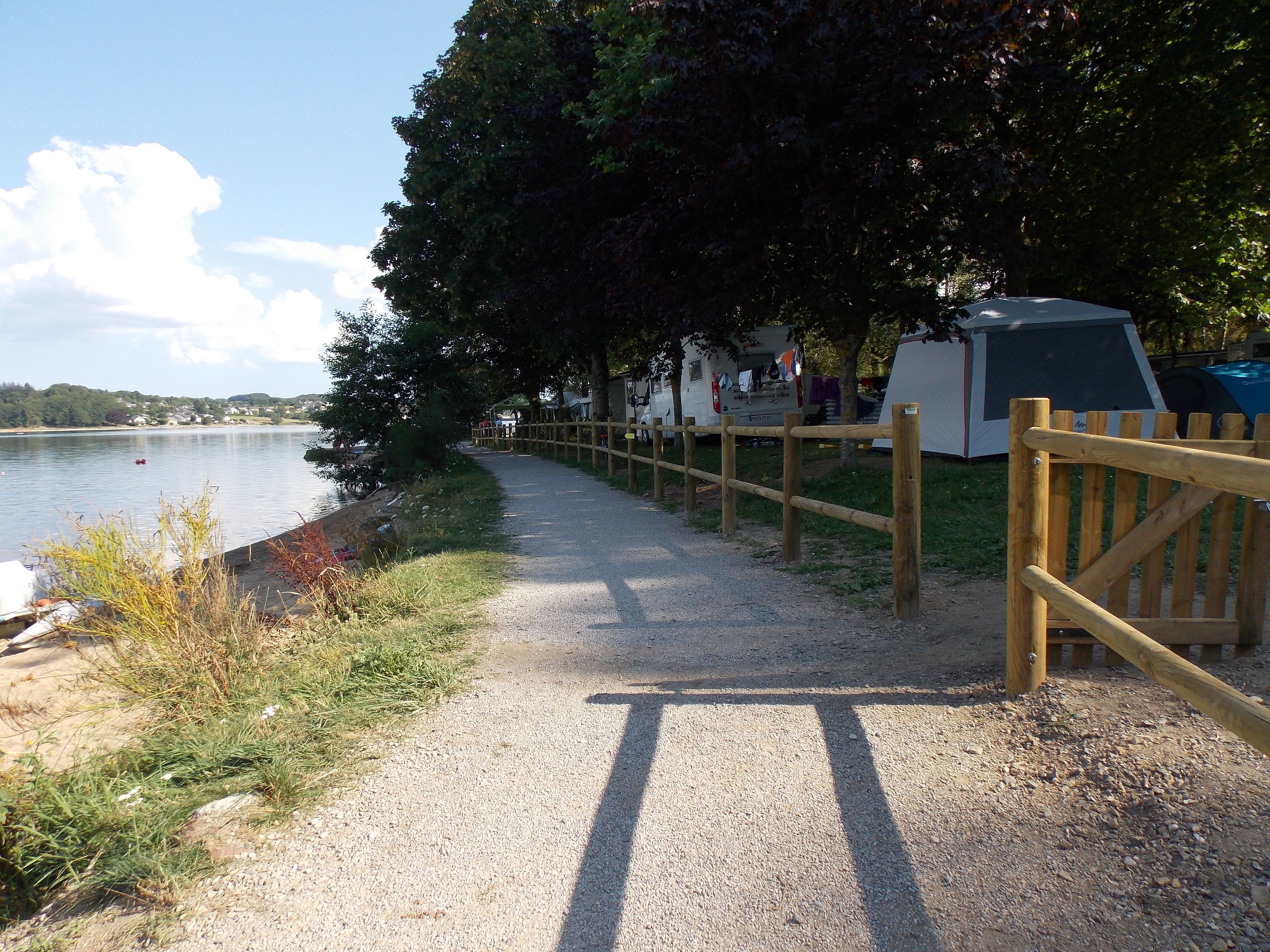 A gravel path beside a lake with a wooden fence, a tent, and a camper parked nearby.