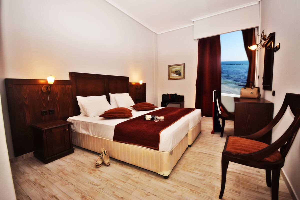 A hotel room with a large bed, wooden headboard, bedside tables, a chair, a desk, and a view of the sea through a window.