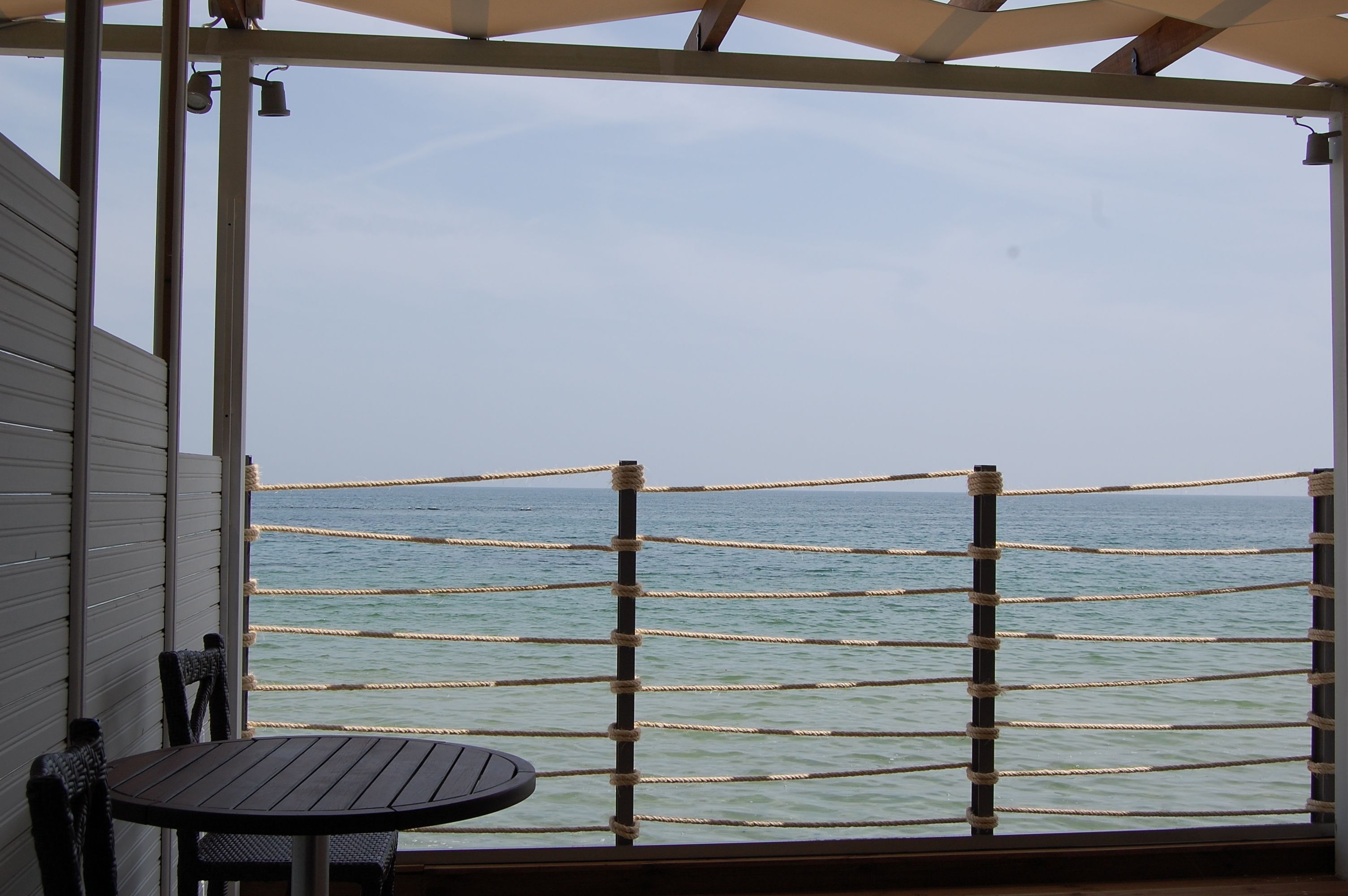 A covered outdoor area with a table and chairs, a rope barrier, and a view of the sea.