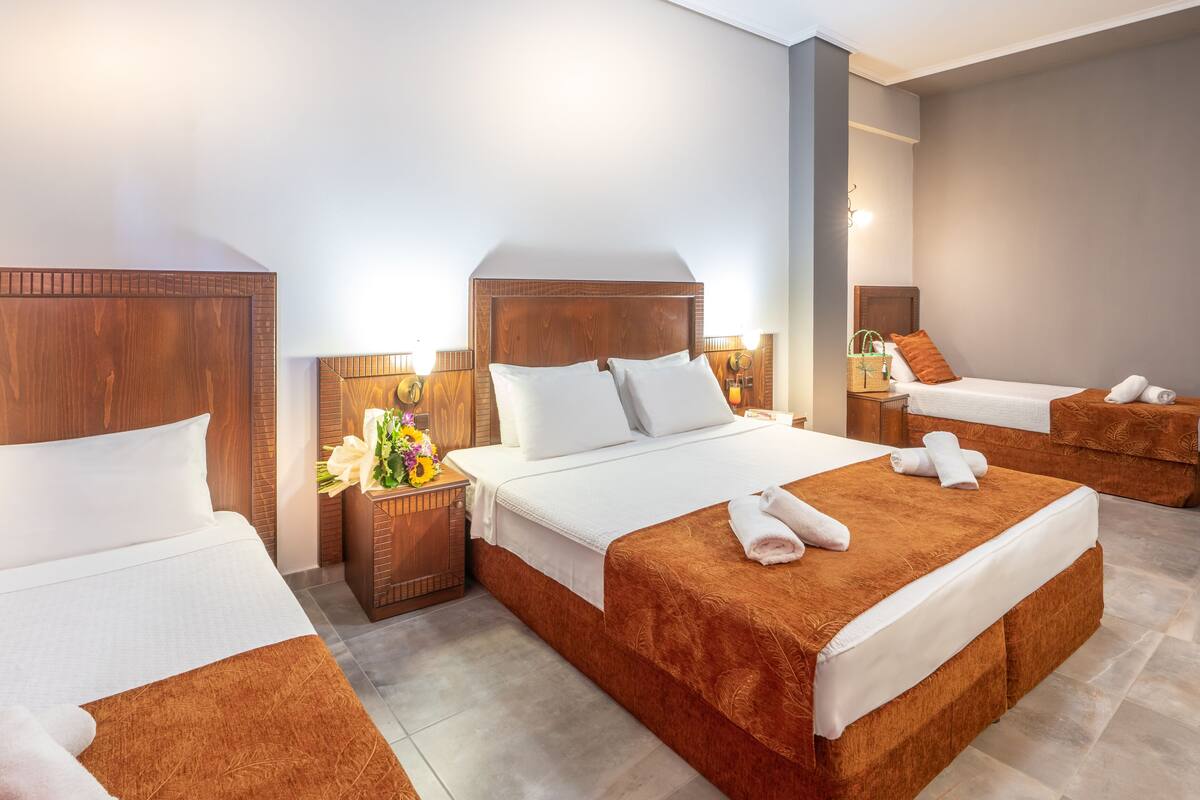 A hotel room with three beds, a wooden headboard, a nightstand with a lamp, and a wall-mounted light fixture.