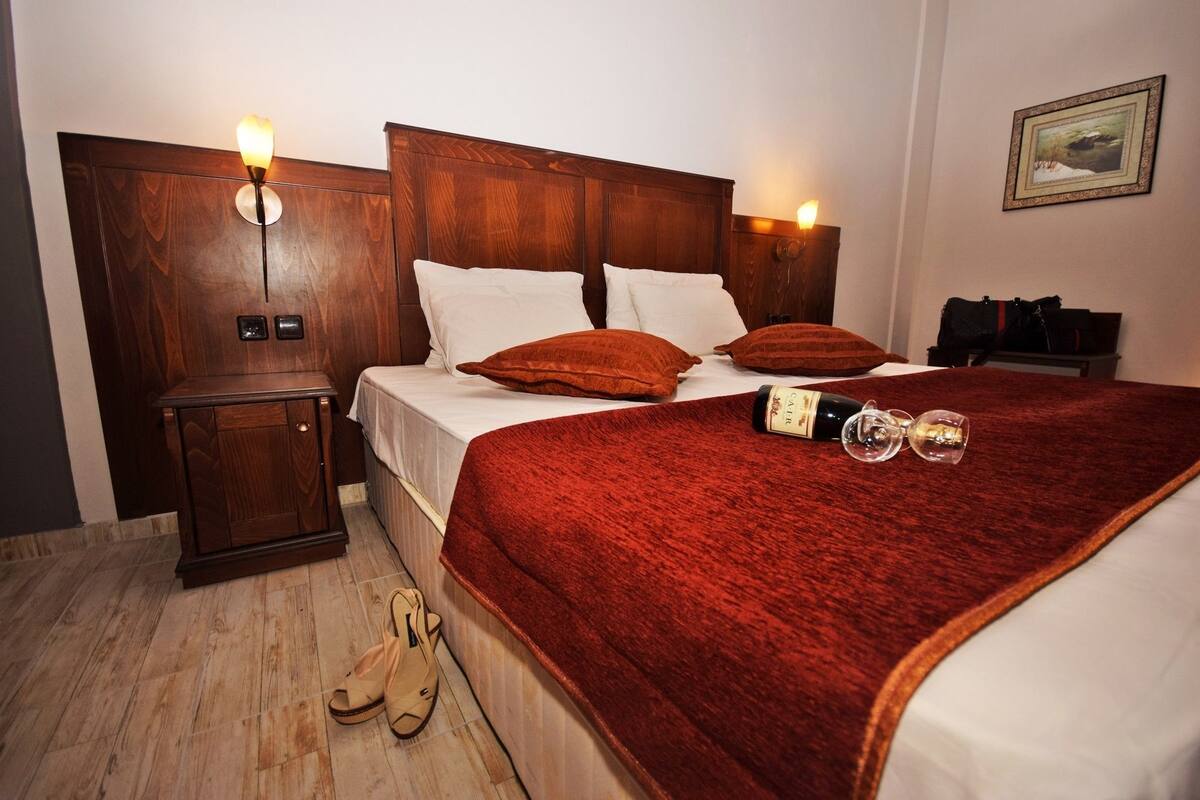 A hotel room with a wooden headboard, two bedside lamps, a bed with white linens and red throw pillows, a red blanket, a bottle of champagne, and a pair of shoes on the floor.