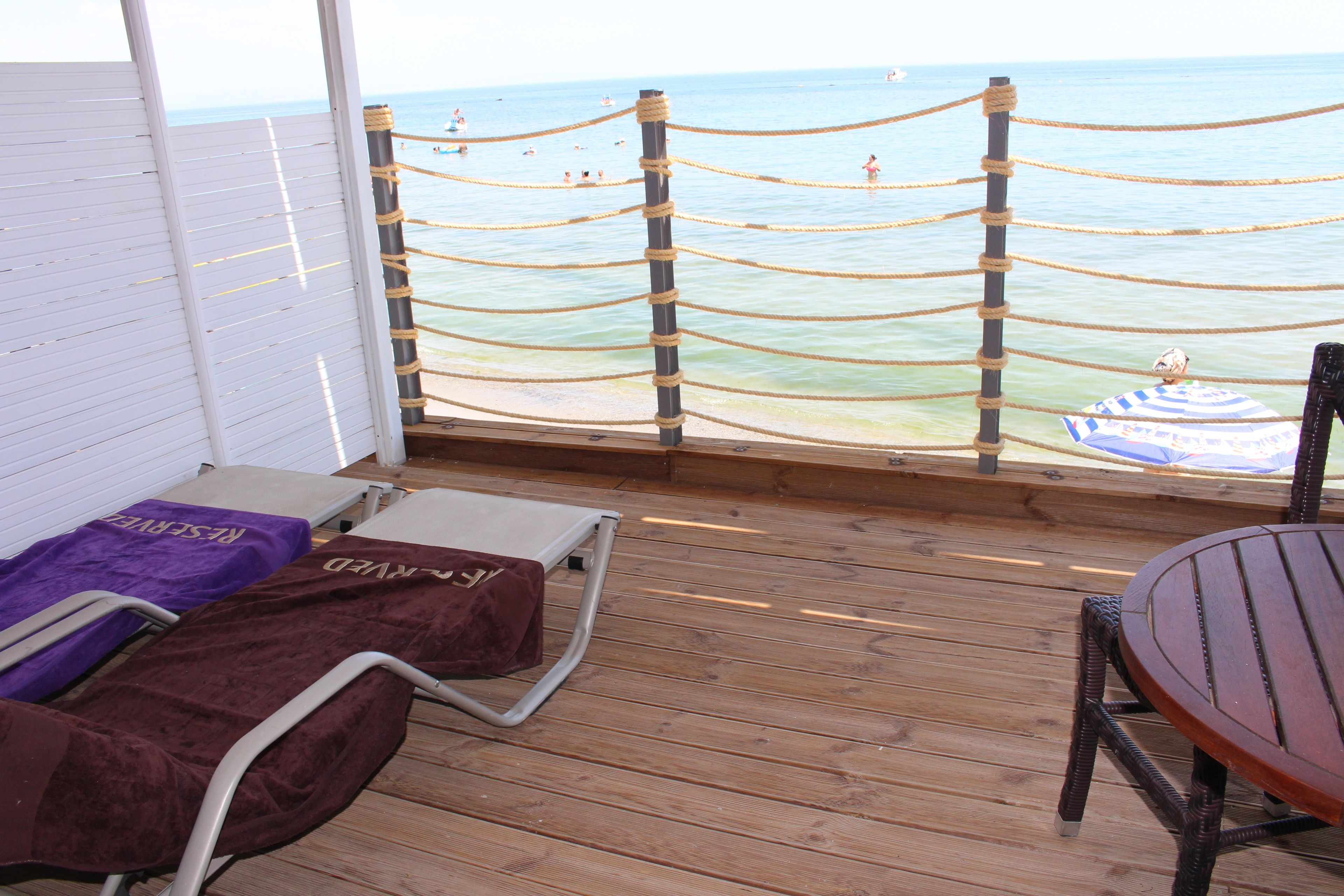 Terraza de madera con sillones de descanso y una mesa, con vista a una playa y una barrera de cuerda.