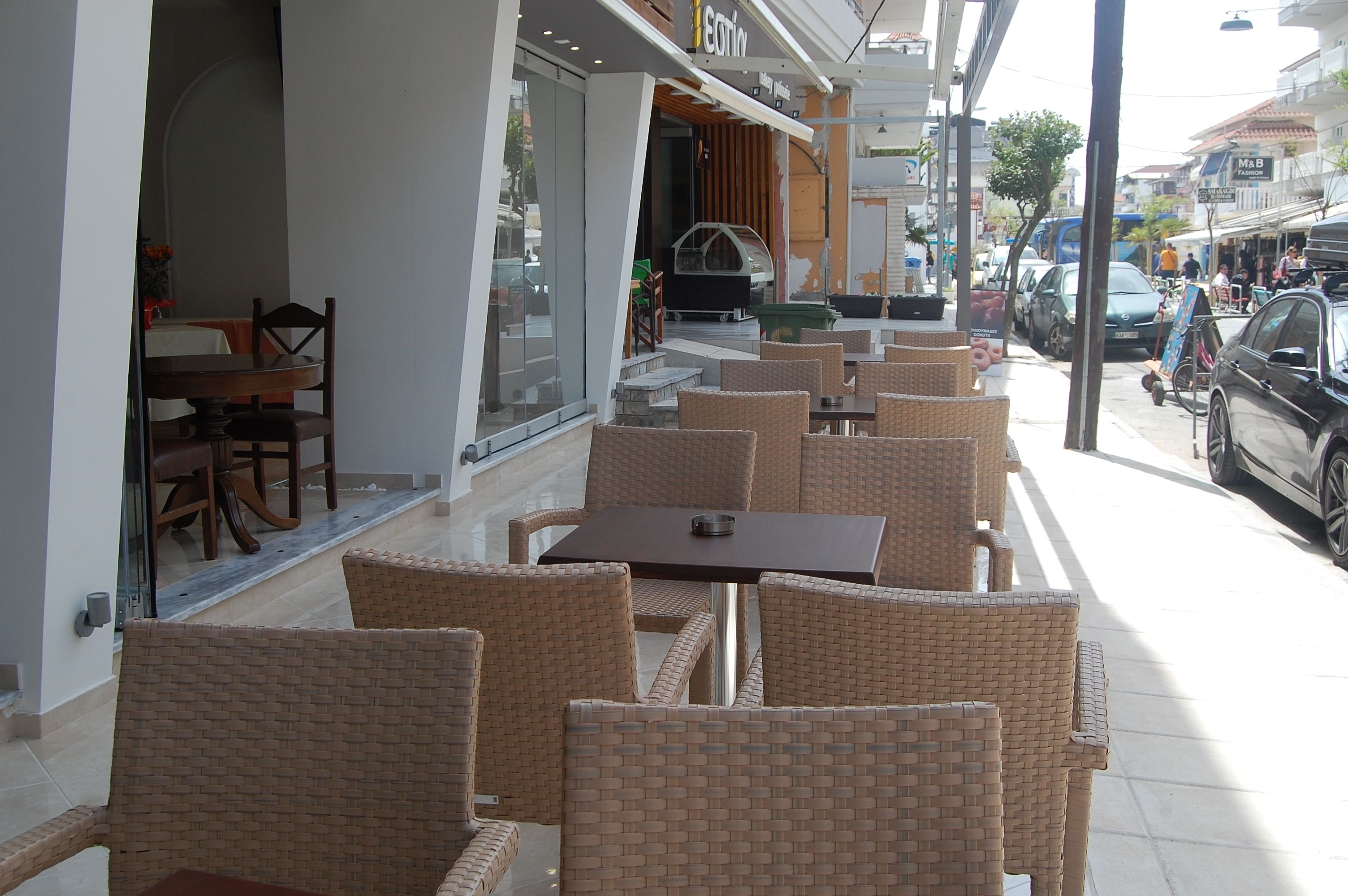An outdoor cafe with wicker chairs and tables, a glass storefront, and a street with parked cars and pedestrians.