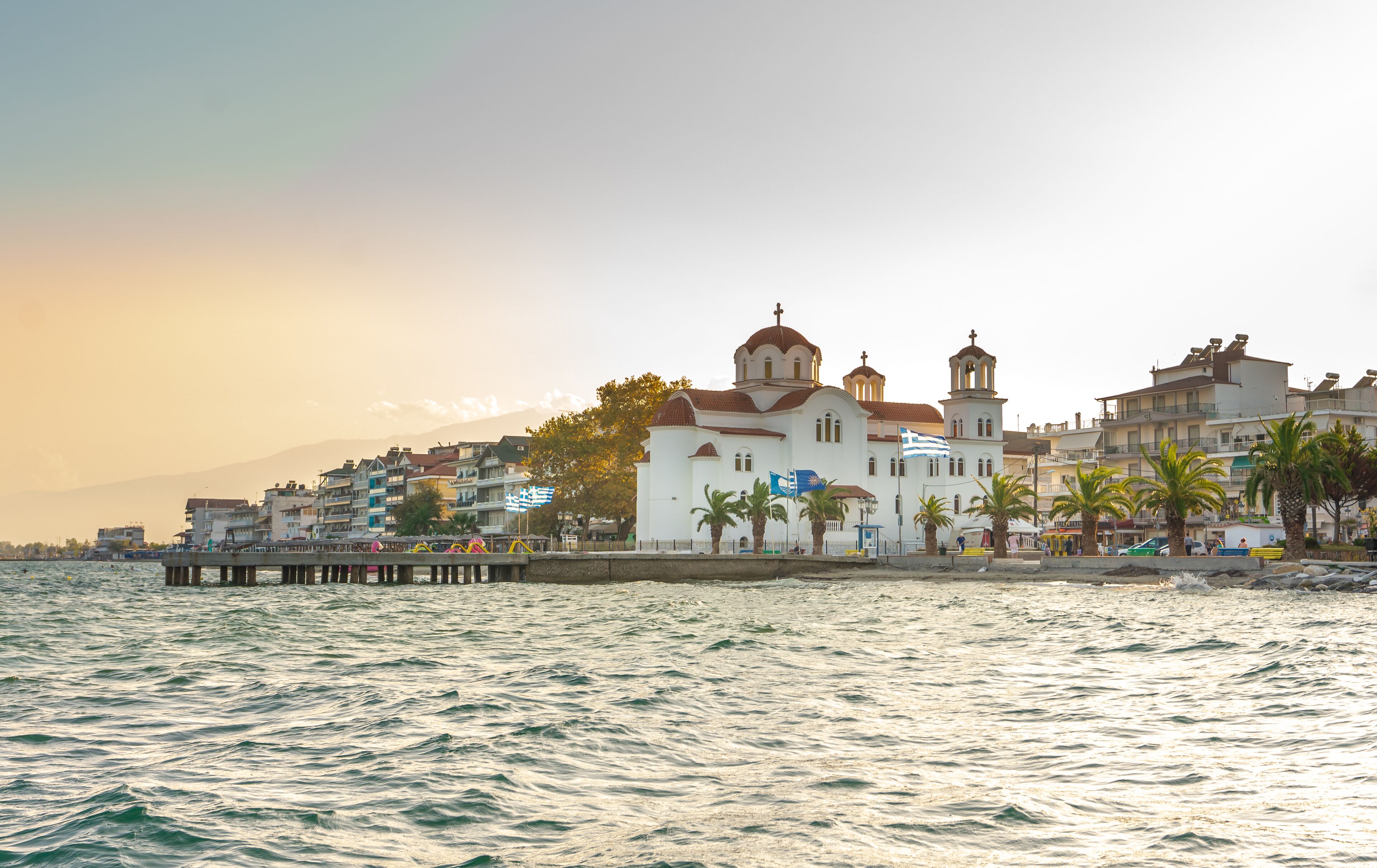 Un pueblo costero con una iglesia blanca destacada con cúpulas, un muelle que se extiende hacia el mar y edificios con balcones.