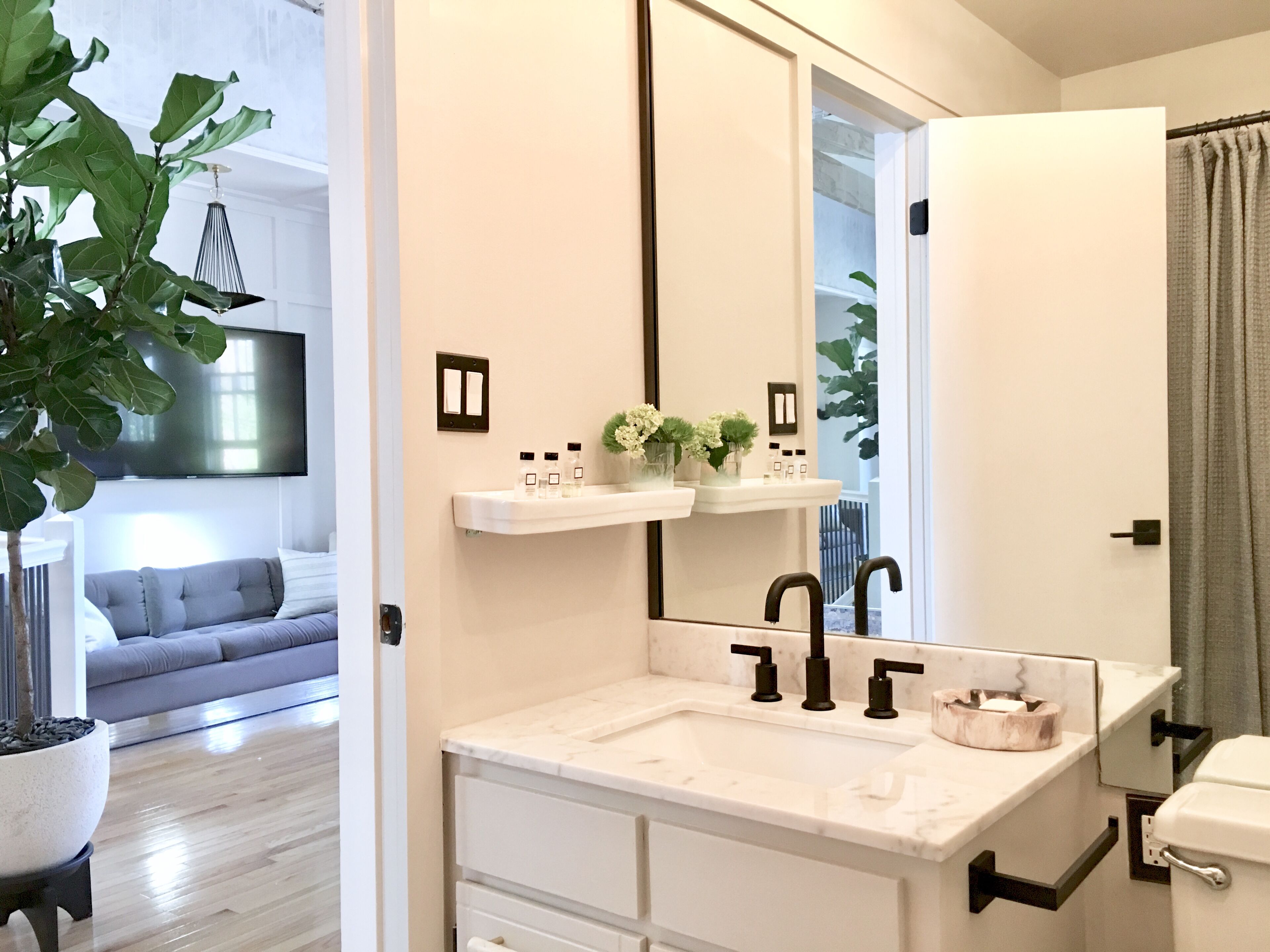 Un baño moderno con un espejo grande, encimera blanca y grifos negros. Hay una planta a la izquierda y un sofá gris al fondo.