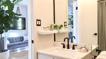Un baño moderno con un espejo grande, encimera blanca y grifos negros. Hay una planta a la izquierda y un sofá gris al fondo.