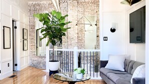 A modern living room with a brick wall, a glass coffee table, a grey sofa, and a large green plant. - Luxuriously Restored Loft in Old Town Alexandria (Alexandria)