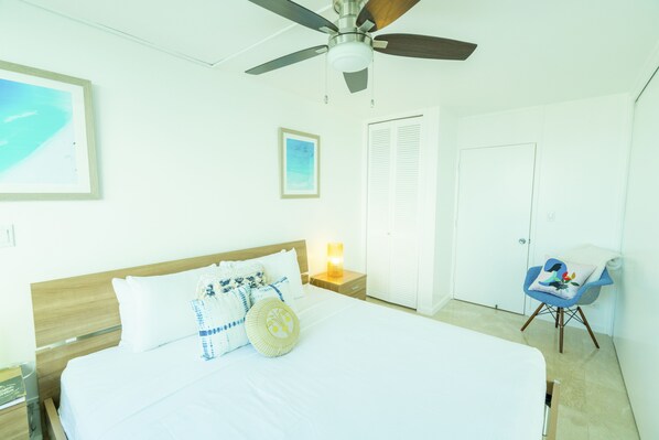 Habitación de hotel con una cama grande, ventilador de techo, mesita de noche con lámpara, una silla y una puerta.