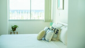 Un dormitorio con un ventanal grande, una cama con ropa de cama blanca y una mesita con una planta.
