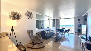 Una sala de estar moderna con un sofá, un sillón mecedora, una mesa de centro y un comedor con sillas y una mesa. Una ventana grande ofrece vistas al océano.