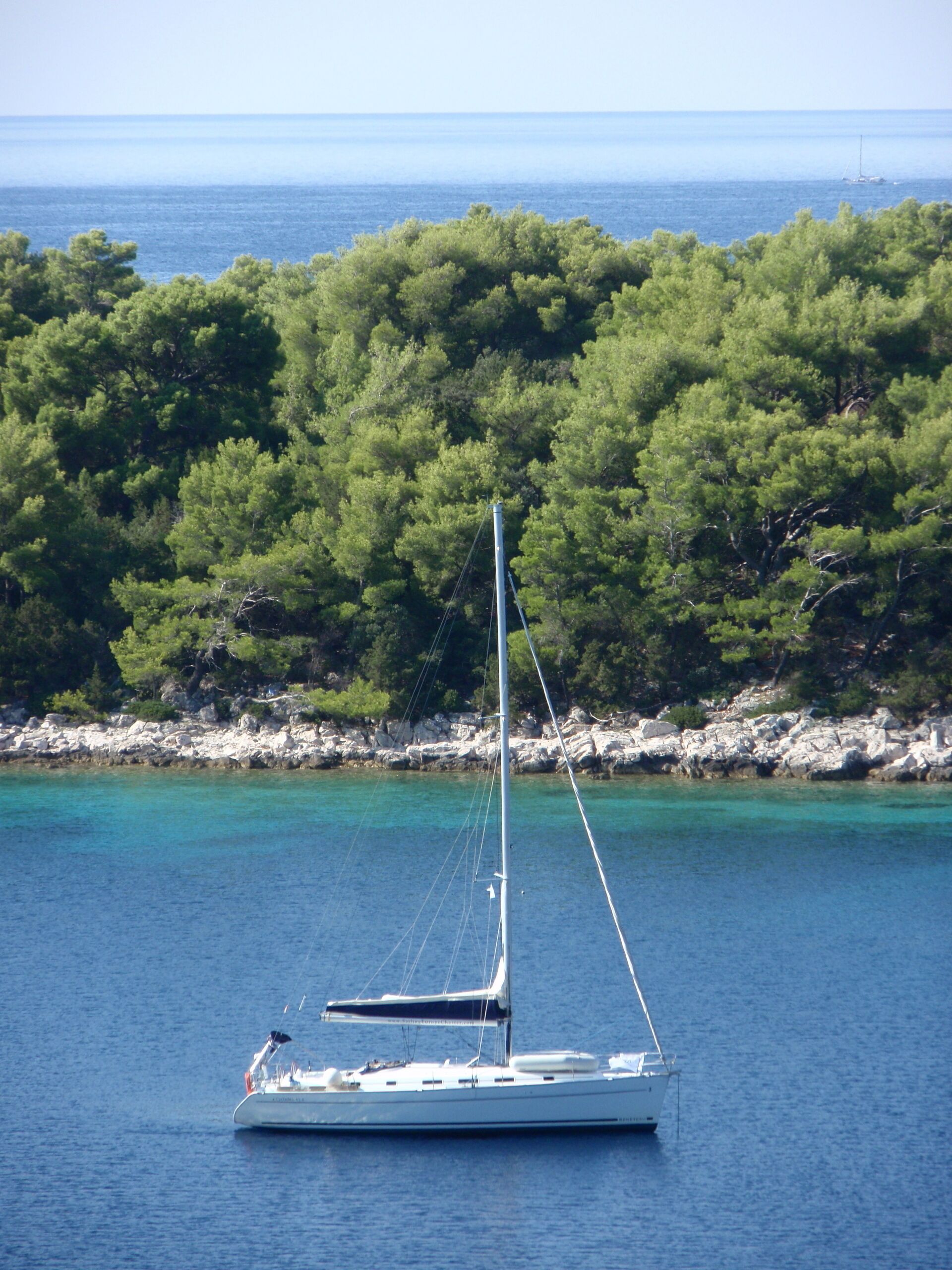 A sailboat on a calm sea with a rocky shoreline and dense green trees in the background.