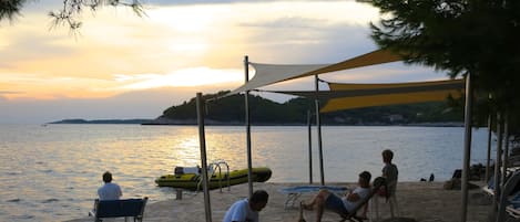 A beach with a stone pavement, a yellow kayak, and people relaxing under a shaded area.