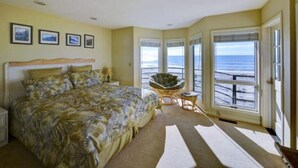 A bedroom with a large bed, a chair, and a view of the beach through large windows. - Luxury Ocean Front Home (Aptos)
