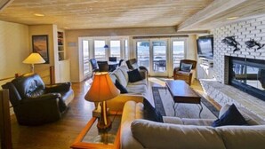 A spacious living room with a wooden ceiling, a fireplace, and large windows offering a view of the sea. - Luxury Ocean Front Home (Aptos)