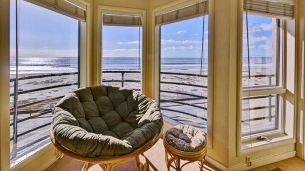 A corner room with large windows overlooking a beach, a round chair, and a small stool. - Luxury Ocean Front Home (Aptos)