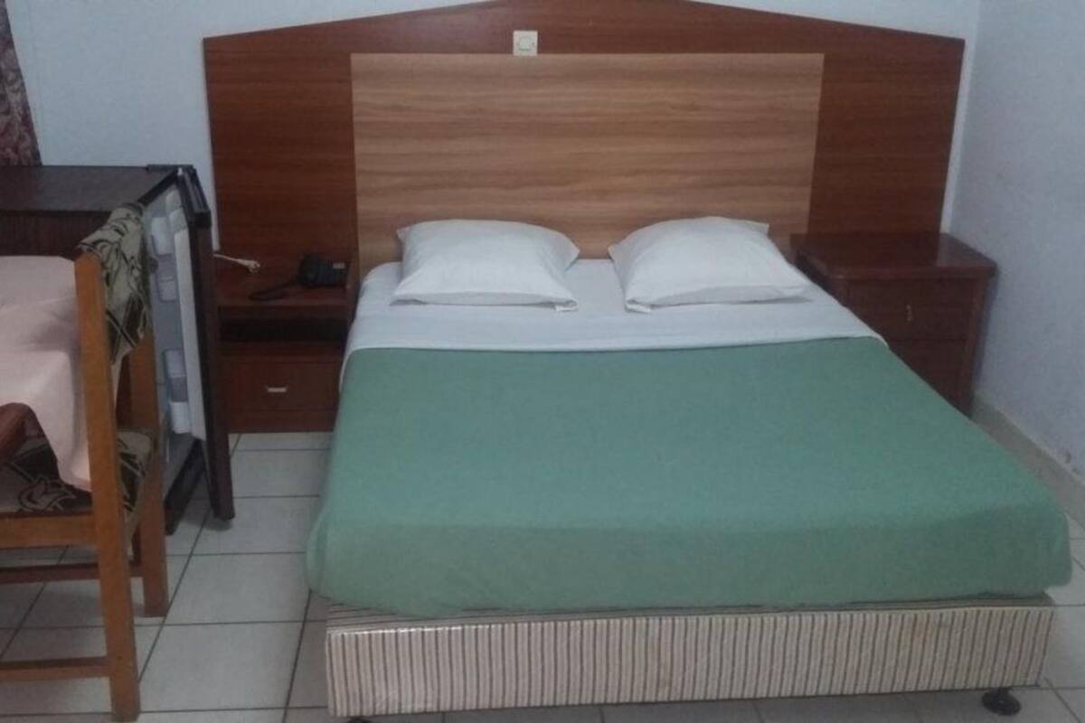 A single bed with a wooden headboard, a green bedspread, and white pillows. There is a wooden nightstand to the left with a lamp and a phone. A wooden chair is positioned at the foot of the bed.