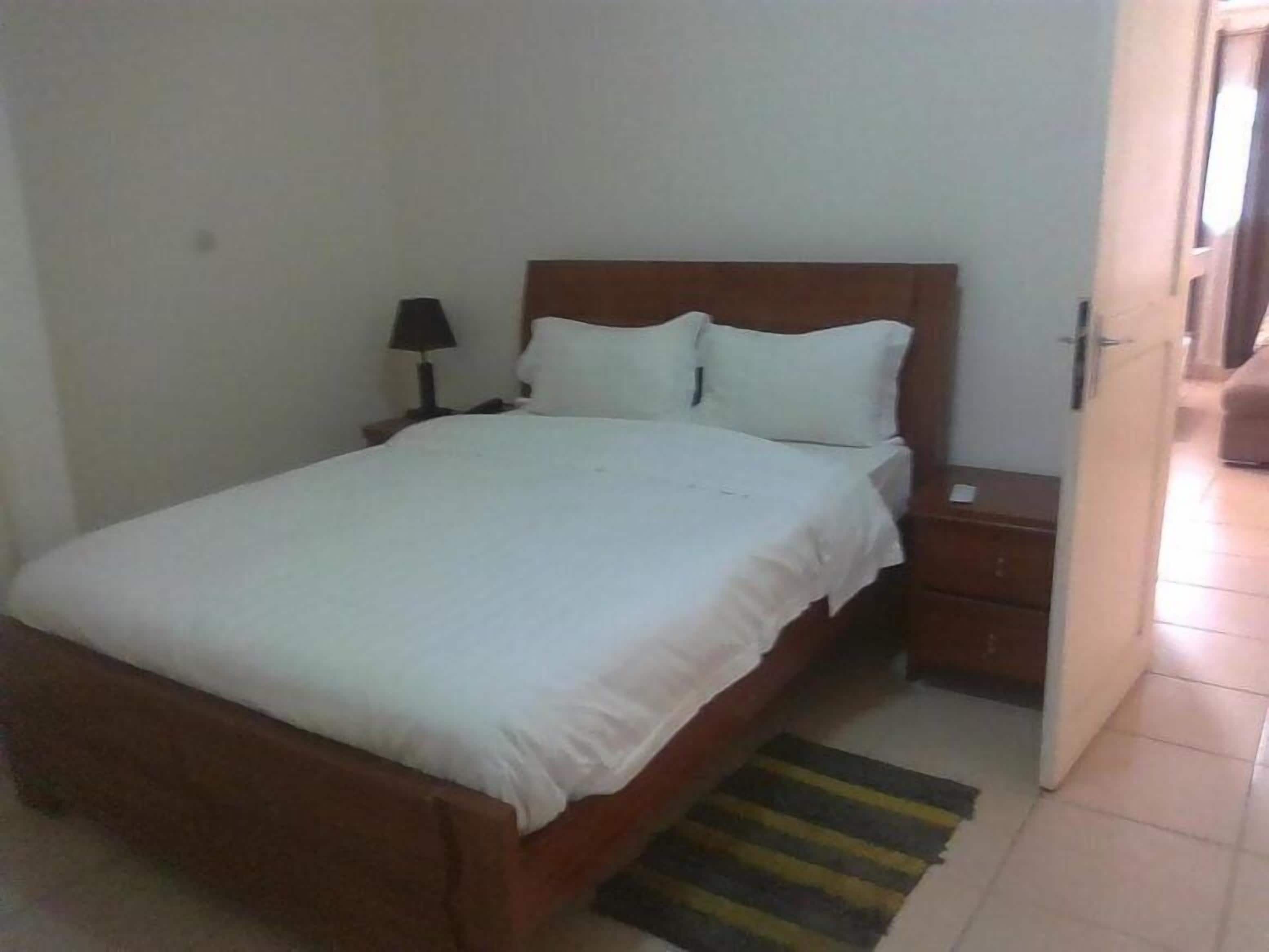 A bedroom with a wooden bed, white bedding, a nightstand with a lamp, and a striped rug.