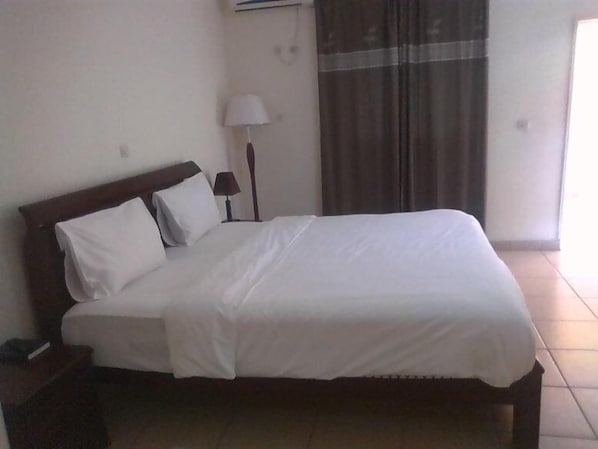A neatly made bed in a hotel room with a wooden headboard, a bedside lamp, and a window with curtains.