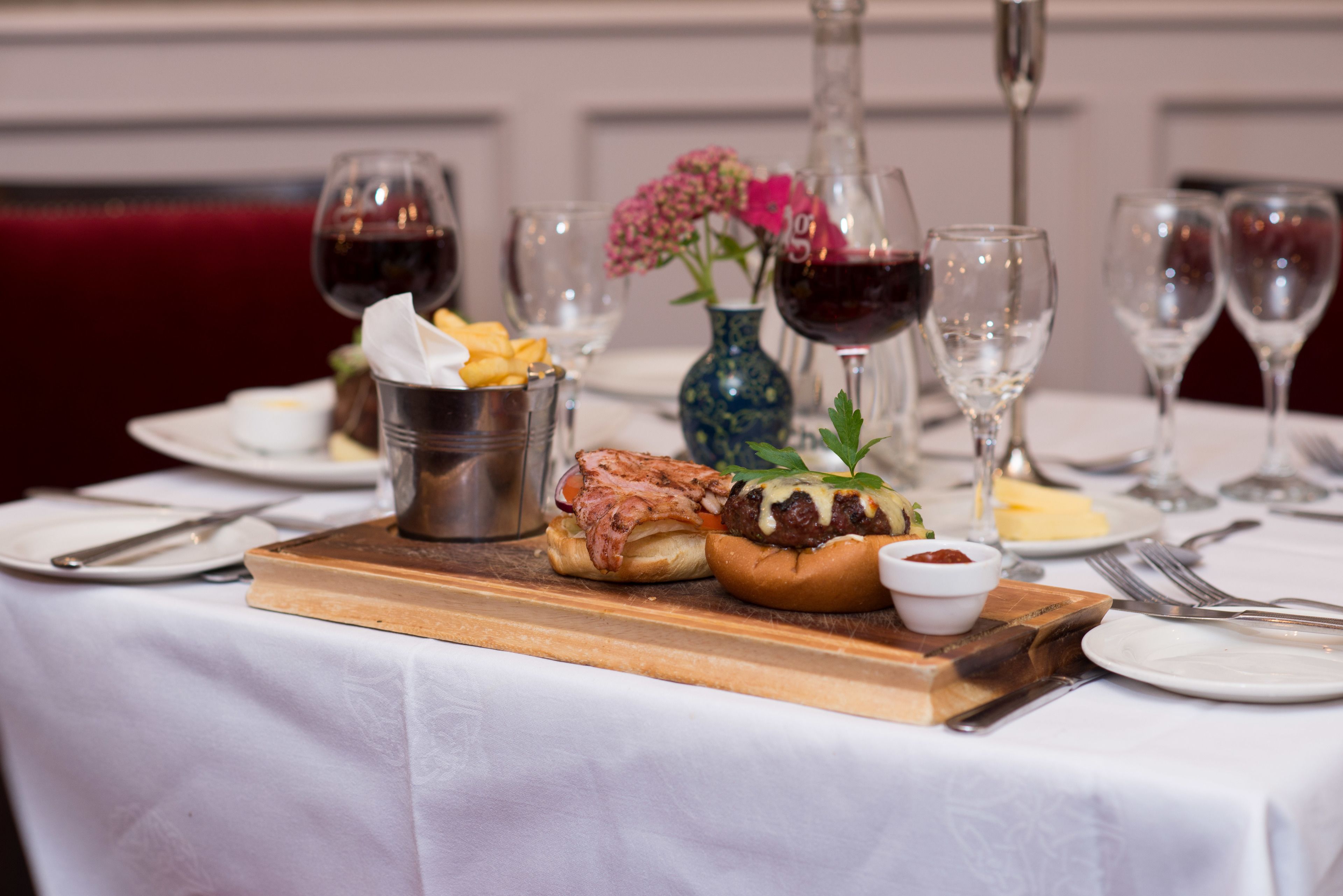 A dining table with a burger, a small bowl of sauce, and a vase with flowers.
