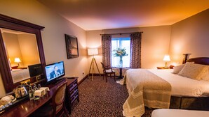 A hotel room with a bed, a desk with a television, a chair, a lamp, and a window with curtains.